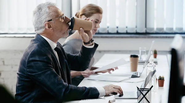 Vue latérale d'un homme d'affaires d'âge moyen buvant du café à emporter et utilisant un ordinateur portable près d'un collègue flou au bureau — Photo de stock