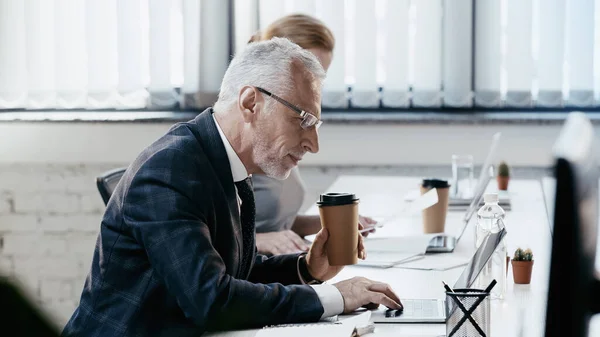 Вид сбоку зрелого бизнесмена, держащего кофе и использующего ноутбук в офисе — стоковое фото