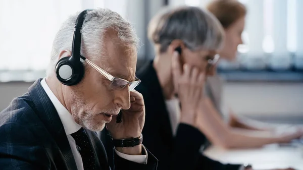 Mature businessman in headset talking near blurred colleagues in office — Stock Photo
