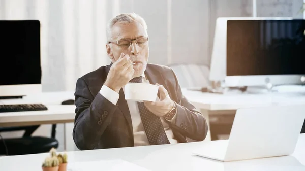 Mature businessman eating takeaway salad near laptop in office — Stock Photo