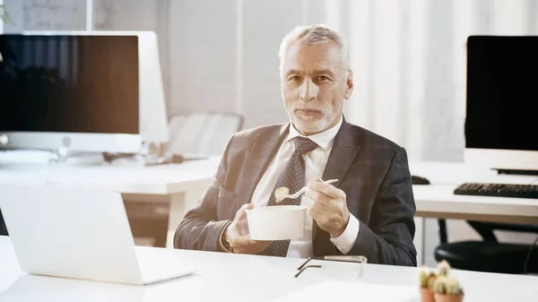 Зрелый бизнесмен смотрит в камеру, держа салат на вынос рядом с ноутбуком в офисе — стоковое фото