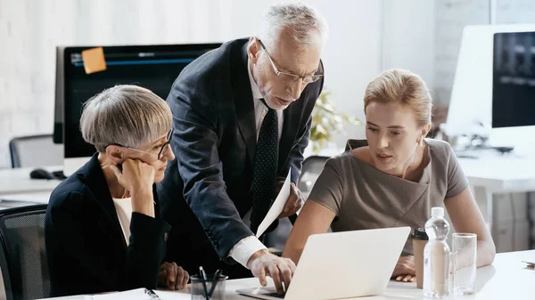 Mature businessman using laptop near colleagues in office — Stock Photo