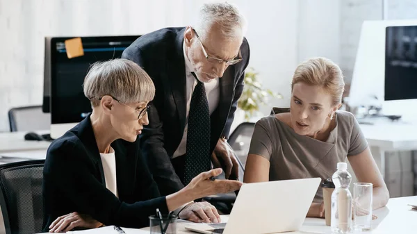 Madura mujer de negocios apuntando a la computadora portátil cerca de colegas en la oficina - foto de stock