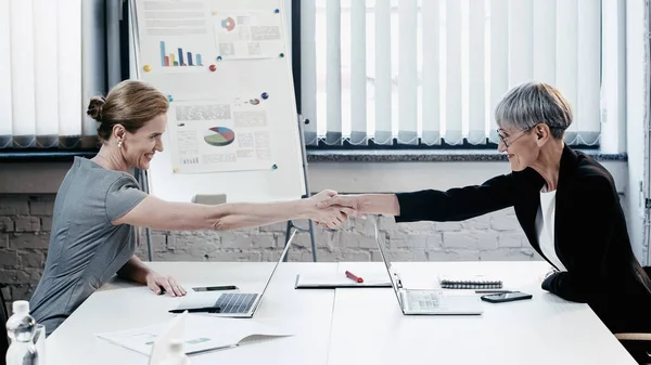 Side view of smiling businesswomen shaking hands near devices in office — Stock Photo