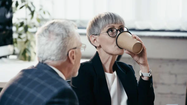 Madura mujer de negocios bebiendo café para ir cerca borrosa colega en la oficina - foto de stock