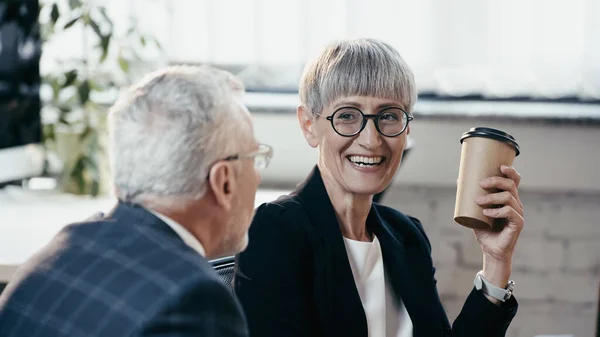 Fröhliche reife Geschäftsfrau hält Kaffee in der Hand, um unscharfen Kollegen im Amt nahe zu kommen — Stockfoto
