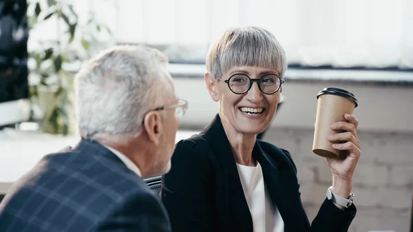 Cheerful mature businesswoman holding coffee to go near blurred colleague in office — Stock Photo