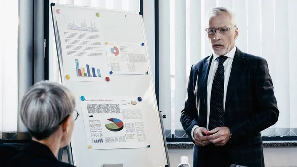 Mature businessman standing near blurred colleague and charts on flip chart in office — Stock Photo