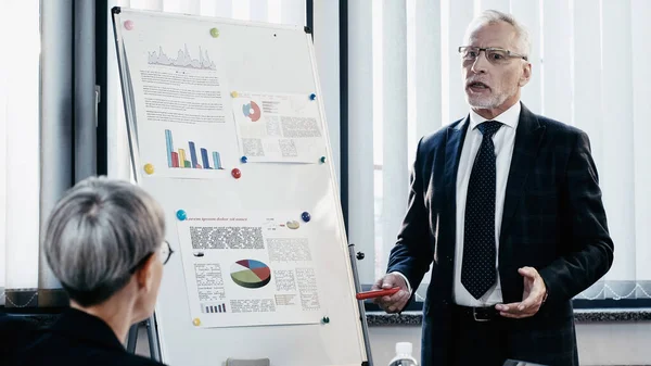 Mature businessman in formal wear talking near flip chart and colleague in office — Stock Photo