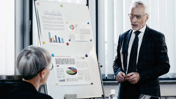 Middle aged businessman talking near blurred businesswoman and flip chart in office — Stock Photo