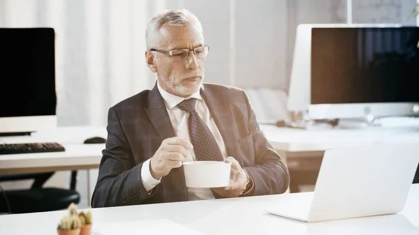 Homme d'affaires d'âge moyen tenant conteneur à emporter près d'un ordinateur portable dans le bureau — Photo de stock