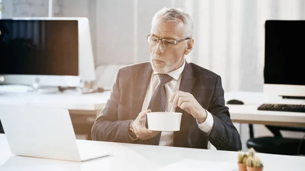 Mature businessman holding takeaway lunch near laptop in office — Stock Photo