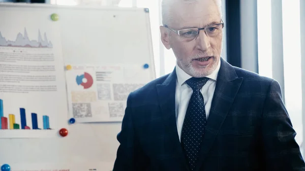 Homme d'affaires mature en tenue formelle et lunettes debout près de flip chart au bureau — Photo de stock