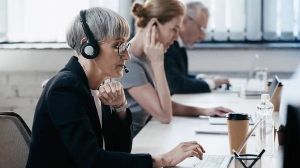 Vue latérale d'une femme d'affaires mature dans un casque à l'aide d'un ordinateur portable près de collègues flous au bureau — Photo de stock