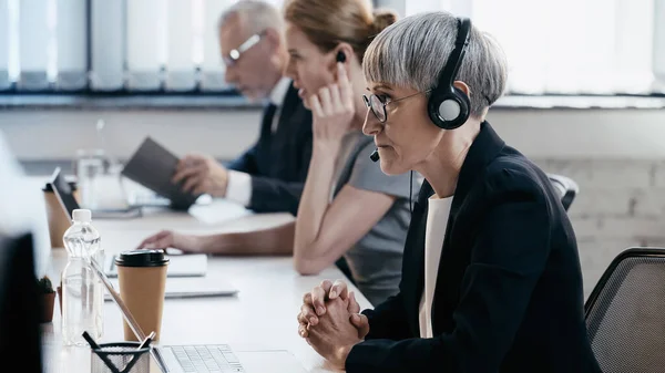 Vista lateral de la mujer de negocios con auriculares cerca de la computadora portátil y bebidas en la oficina - foto de stock