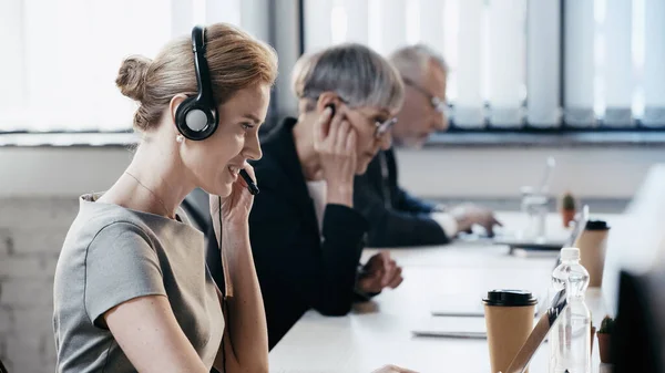 Vue latérale d'une femme d'affaires souriante utilisant un casque près d'un ordinateur portable et un café pour aller au bureau — Photo de stock