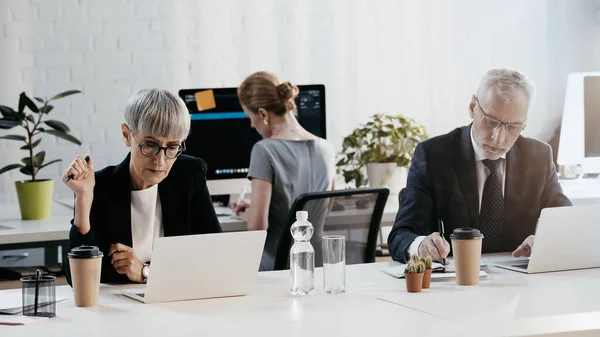 Donna d'affari matura guardando computer portatile vicino a bevande e collega in carica — Foto stock