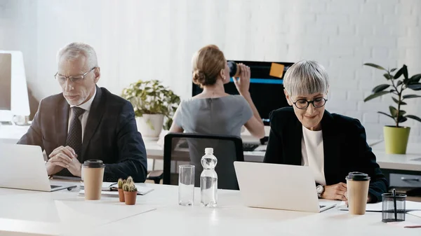 Усміхнена бізнес-леді дивиться на папери біля кави, щоб піти і ноутбук в офісі — стокове фото