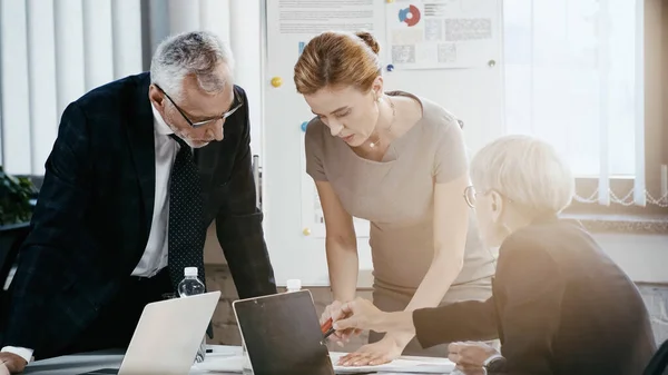 Gente de negocios mirando documentos cerca de colega apuntando a la computadora portátil en la oficina - foto de stock