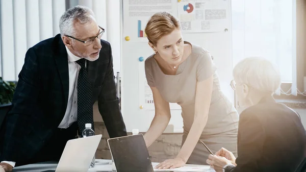 Gente de negocios mirando a su colega mientras trabaja cerca de computadoras portátiles en la oficina - foto de stock