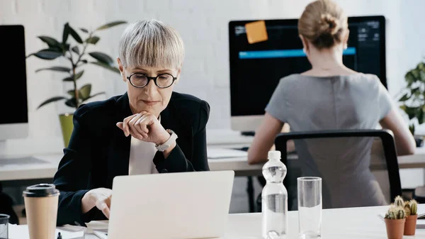 Зріла бізнес-леді в окулярах використовує ноутбук біля паперової чашки в офісі — стокове фото
