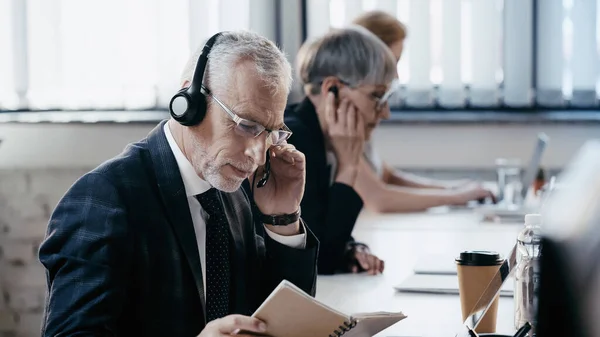 Hombre de negocios maduro en auriculares mirando portátil cerca de la computadora portátil y café en la oficina - foto de stock