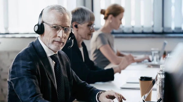 Homme d'affaires mature dans un casque regardant la caméra près d'un ordinateur portable et du café pour aller au bureau — Photo de stock