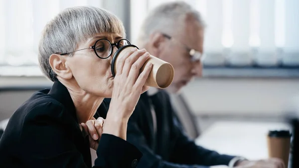 Mature businesswoman in eyeglasses drinking coffee to go near blurred businessman in office — Stock Photo