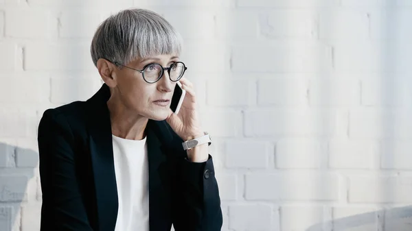 Mature businesswoman in eyeglasses talking on smartphone in office — Stock Photo