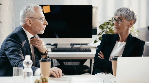 Зрелый бизнесмен разговаривает с коллегой возле бумажных стаканчиков и ноутбука в офисе — стоковое фото