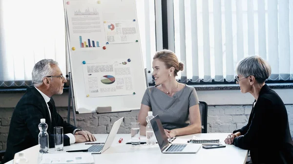 Улыбающаяся деловая женщина разговаривает со зрелыми коллегами возле бумаг и устройств в офисе — стоковое фото