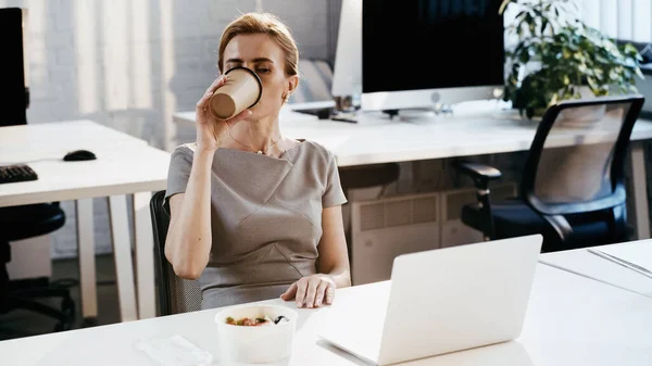 Businesswoman drinking coffee to go near takeaway salad and laptop in office — Stock Photo