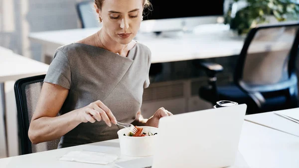 Mujer de negocios sentada cerca de ensalada para llevar y computadora portátil en la oficina - foto de stock