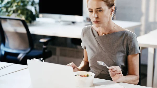 Businesswoman holding fork near takeaway salad and laptop in office — Stock Photo