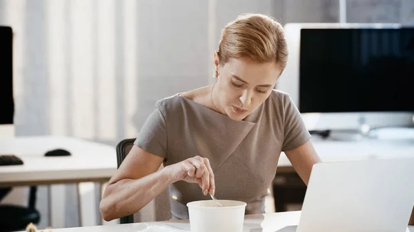 Femme d'affaires tenant fourchette près de emporter déjeuner et ordinateur portable dans le bureau — Photo de stock