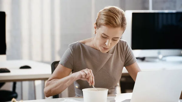 Businesswoman holding fork near takeaway lunch and laptop in office — Stock Photo