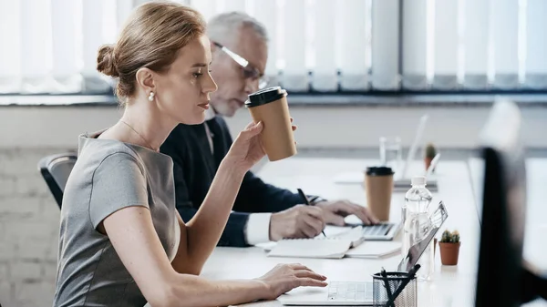 Vista laterale della donna d'affari che tiene la tazza di carta e utilizza il computer portatile in ufficio — Foto stock