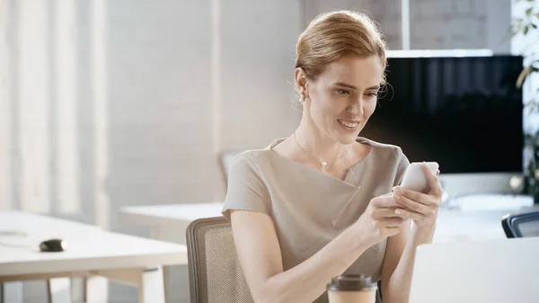 Улыбающаяся деловая женщина, использующая смартфон рядом с размытым ноутбуком и кофе, чтобы пойти в офис — стоковое фото