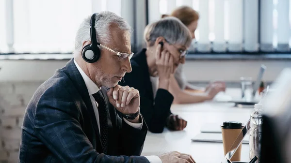 Uomo d'affari di mezza età in cuffia utilizzando laptop vicino a bevande in ufficio — Foto stock