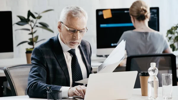 Homme d'affaires mature utilisant un ordinateur portable et détenant des documents près des appareils dans le bureau — Photo de stock