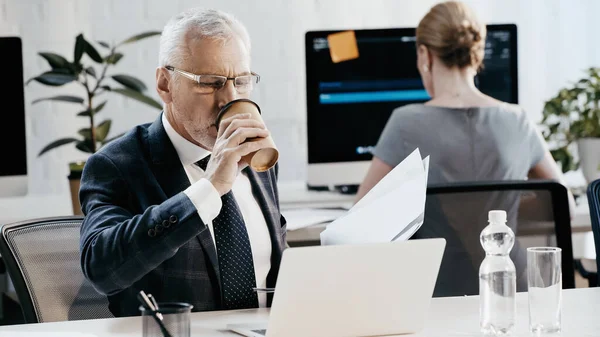 Uomo d'affari maturo che beve caffè per andare a tenere documenti vicino al computer portatile in ufficio — Foto stock