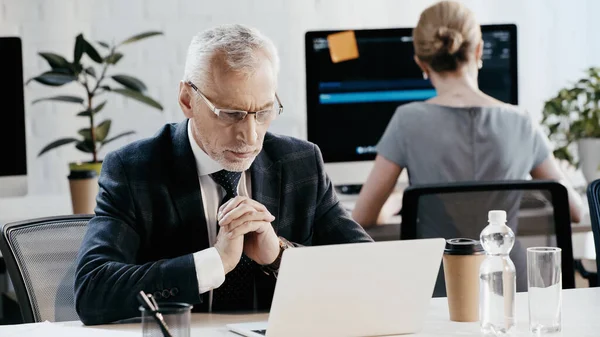 Businessman in eyeglasses looking at laptop near drinks in office — Stock Photo