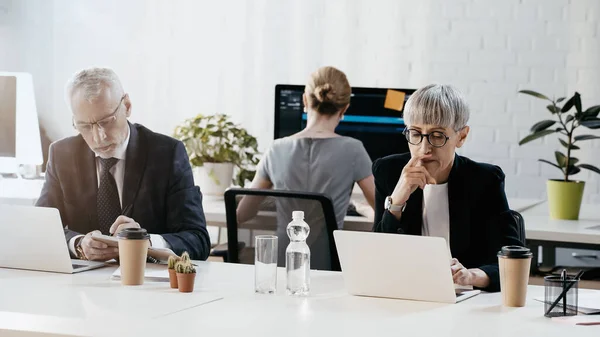 Donna d'affari che lavora su computer portatile vicino a bevande e collega con notebook in ufficio — Foto stock