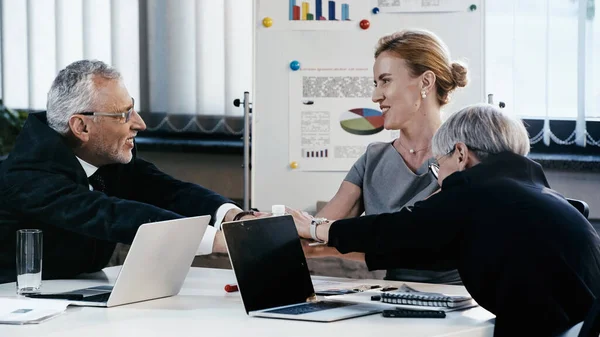 Alegre gente de negocios tomados de la mano cerca de gadgets durante la reunión en la oficina - foto de stock