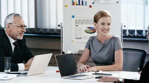 Gente alegre de negocios hablando con colegas cerca de computadoras portátiles y rotafolio en la oficina - foto de stock