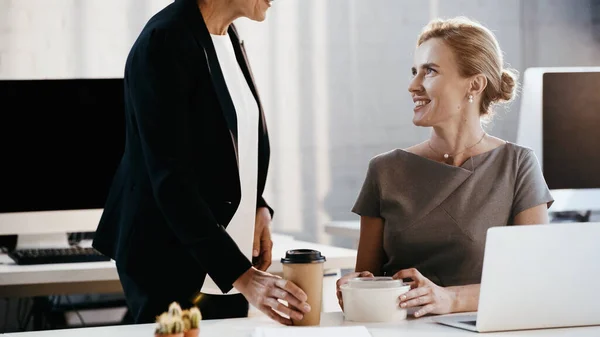 Smiling businesswoman holding takeaway lunch near colleague with coffee to go in office — Stock Photo