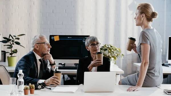Empresária segurando café para ir enquanto conversa com colegas perto de laptop e papéis no escritório — Stock Photo