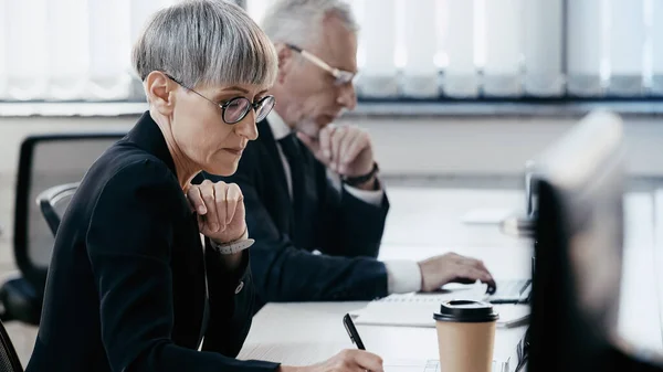 Donna d'affari di mezza età che tiene la penna vicino al laptop e alla tazza di carta in ufficio — Foto stock