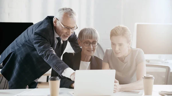 Lächelnder Geschäftsmann mit Brille, Laptop in der Nähe von Kollegen und Coffee to go im Büro — Stockfoto