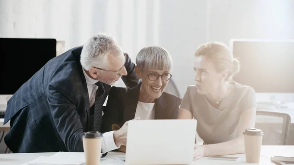 Cheerful business people looking at colleague near devices and coffee in office — Stock Photo